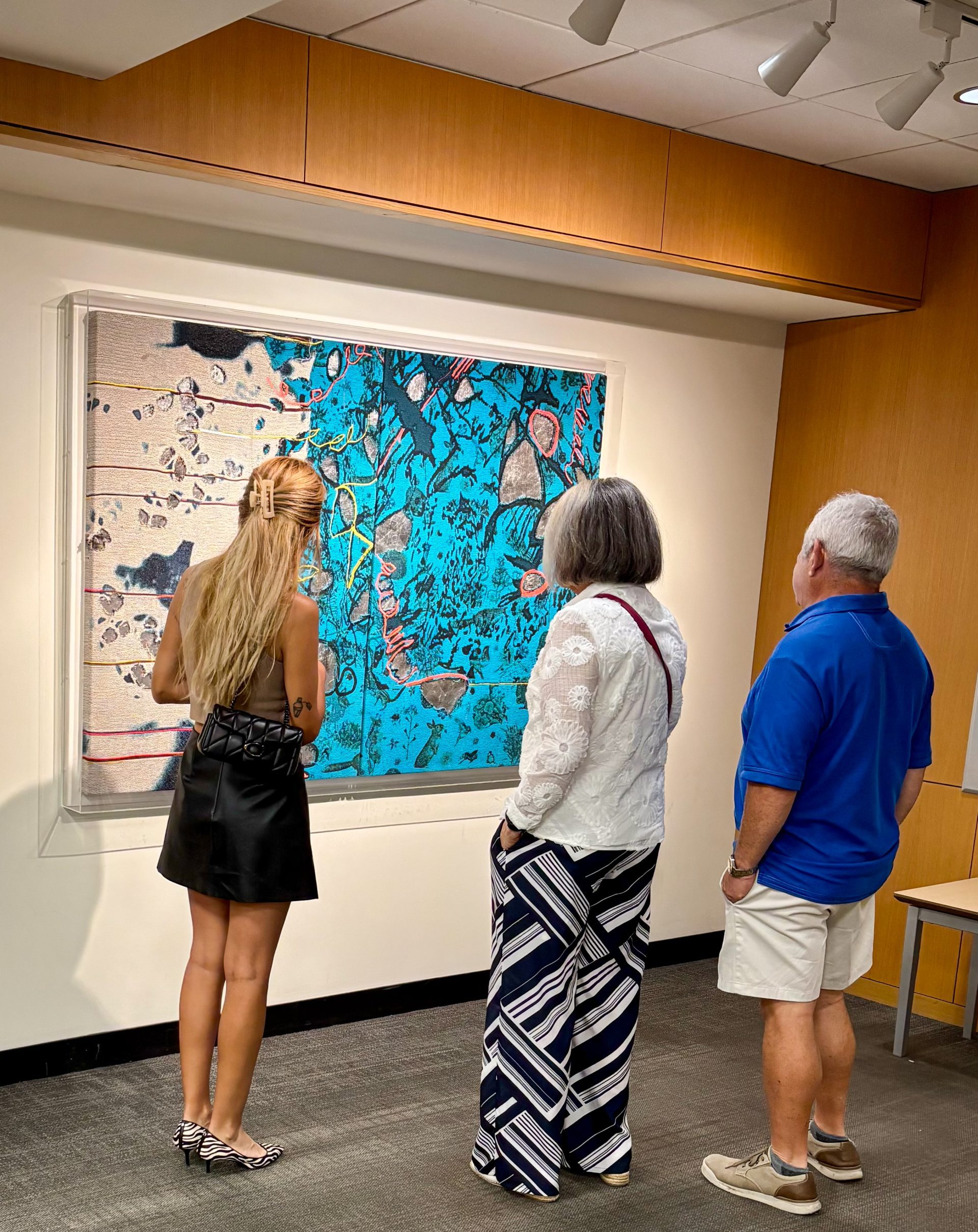 Three people are viewing Melissa Leandro's Aqua Melt/Flores y Conejos artwork, a large abstract textile piece, in the DGPL lobby